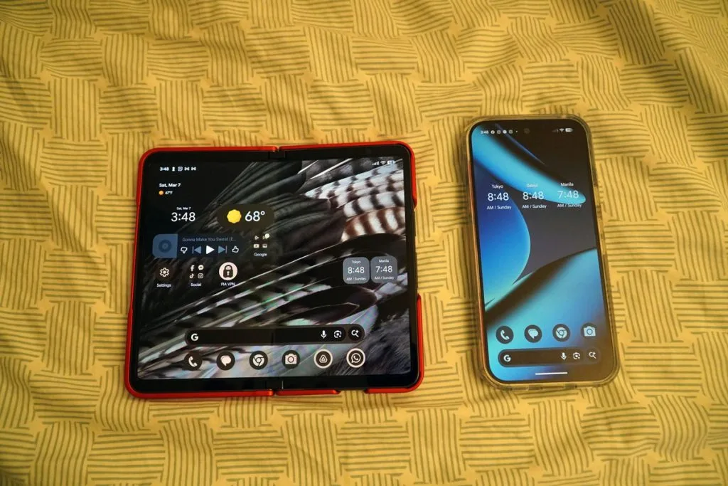 Google Pixel Fold unfolded in red case next to a standard smartphone on a patterned yellow fabric.