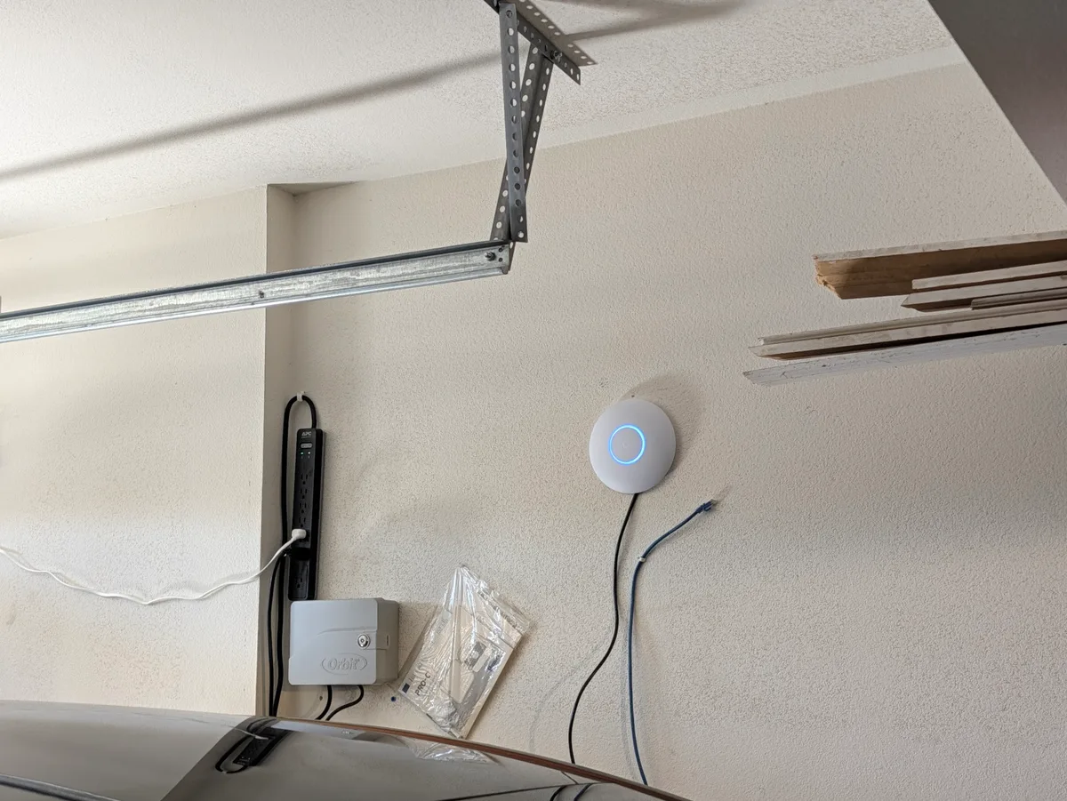 Wider view of the garage wall showing the U7 Lite alongside the power strip and sprinkler controller